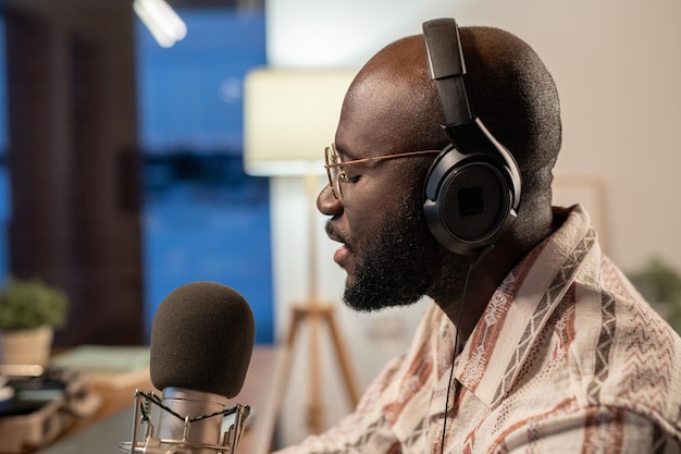 Zijaanzicht van jonge zwarte man in koptelefoon en bril zingen in studio