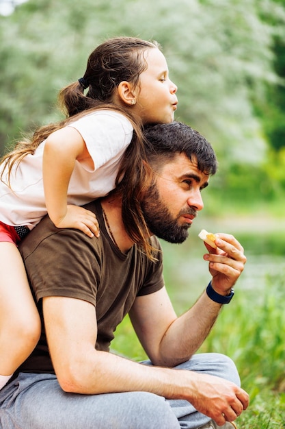 Zijaanzicht van lachende familie zittend op picknick in parkbos rond bomen Dochtertje zittend op vaders rug