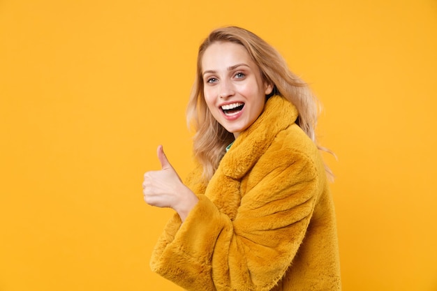 Zijaanzicht van lachende jonge blonde vrouw meisje in gele bontjas poseren geïsoleerd op een oranje achtergrond, studio portret. Mensen levensstijl concept. Bespotten kopie ruimte. Het tonen van duim omhoog kijkende camera.