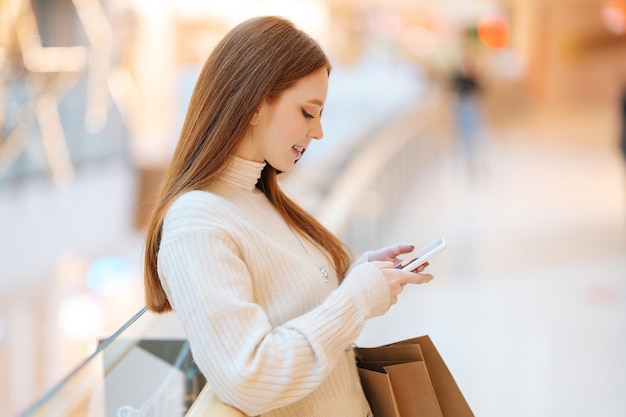 Zijaanzicht van lachende roodharige jonge vrouw met behulp van smartphone winkelen papieren zakken met aankoop staande in de hal van winkelcentrum met licht interieur kijken op scherm wazig achtergrond