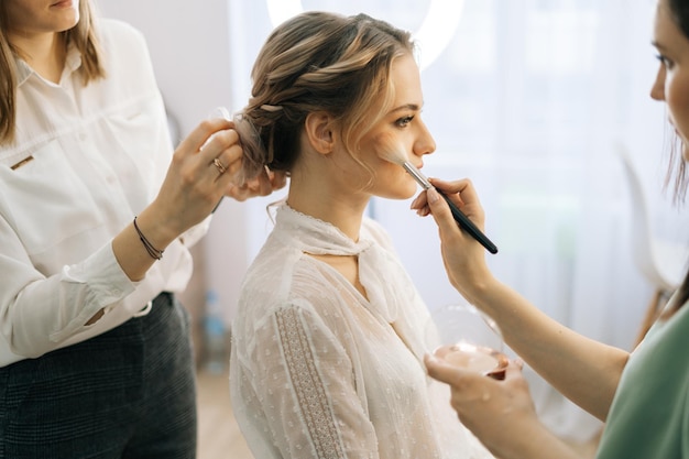 Zijaanzicht van make-up artiest die rouge op de wang van de vrouw zet met borstel terwijl kapper kapsel maakt voor jonge vrouw in schoonheidssalon. Concept van backstage werk.