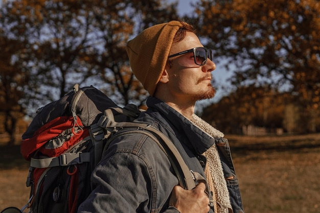 Foto zijaanzicht van mannelijke reiziger met wandelrugzak in zonnebril en hipster-slijtage staande op zonnige dag