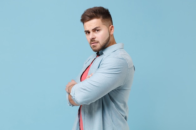 Zijaanzicht van ontevreden jonge bebaarde man 20s in casual shirt poseren geïsoleerd op pastel blauwe achtergrond in studio. Mensen levensstijl concept. Bespotten kopie ruimte. Hand in hand gekruist uitziende camera.