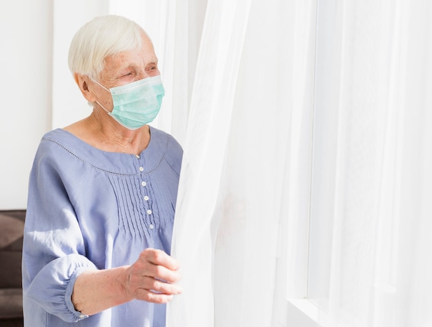 Foto zijaanzicht van oudere vrouw met medisch masker dat door het venster kijkt