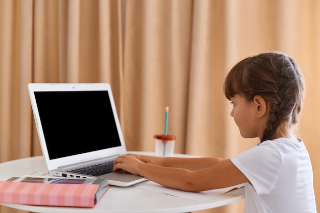 Zijaanzicht van schattig charmant klein meisje met vlechten met online les tijdens afstandsonderwijs zittend op tafel en typen online taak voor school