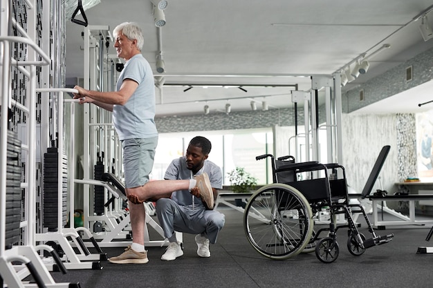 Zijaanzicht van senior man die rehabilitatieoefeningen doet in de sportschool met therapeut die kopieerruimte helpt