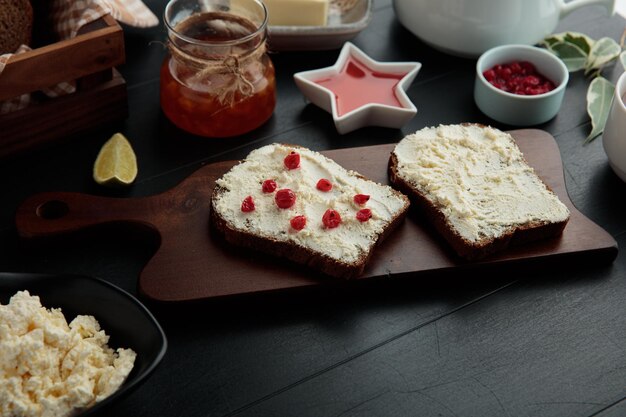 Zijaanzicht van sneetjes brood besmeurd met kwark met rode aalbessen erop op snijplank met jam boter limoen schijfje rode bes op zwarte achtergrond