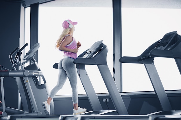 Zijaanzicht van sportieve blonde vrouw in roze pet uitoefenen op loopband in de sportschool