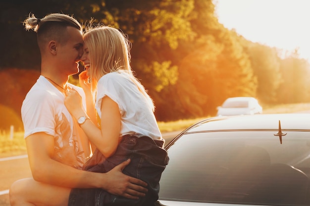 Zijaanzicht van stijlvolle knappe jongeman omarmen prachtige blonde vrouw zittend op de auto kofferbak beide lachen om zonsondergang op backlit van de weg