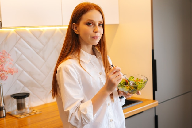 Zijaanzicht van vrij aantrekkelijke jonge roodharige vrouw die verse vegetarische salade eet die van vers geniet