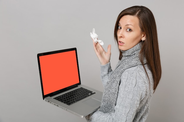 Zijaanzicht van vrouw in trui sjaal met papieren servet met laptop pc-computer met leeg leeg scherm geïsoleerd op een grijze achtergrond. Gezonde levensstijl online behandeling die het concept van het koude seizoen raadpleegt.