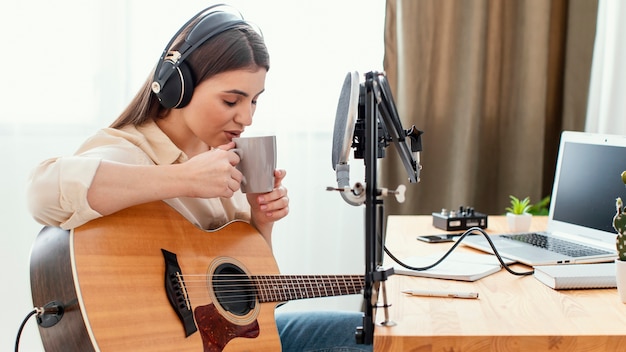 Foto zijaanzicht van vrouwelijke muzikant met een drankje tijdens het thuis spelen van akoestische gitaar