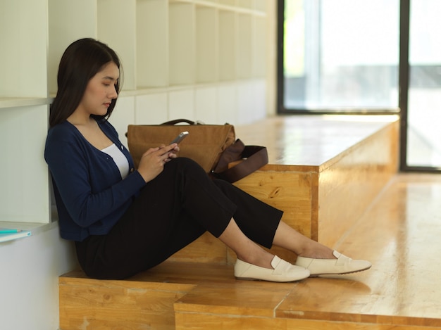 Zijaanzicht van vrouwelijke universiteitsstudent die smartphone gebruikt terwijl ontspannen in co werkruimte zit
