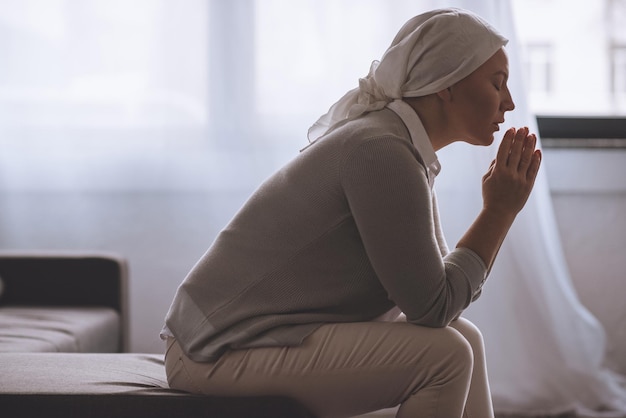 Zijaanzicht van zieke volwassen vrouw in hoofddoek bidden, kankerconcept