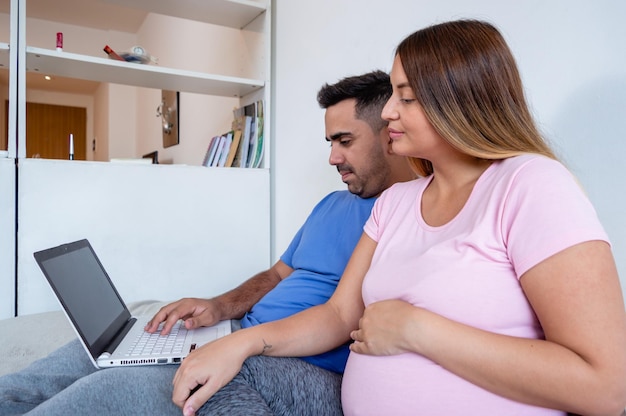 zijaanzicht van zwangere vrouw met haar man zittend op het bed kijkend naar het scherm van de laptop