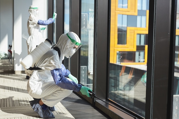 Zijaanzicht volledige lengte portret van twee vrouwelijke werknemers dragen hazmat pakken desinfecteren ramen in kantoorgebouw,