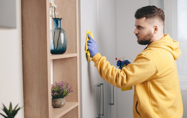 Zijaanzichtfoto van een blanke man met baard en gele hoodie die het huis schoonmaakt met handschoenen en speciale spray