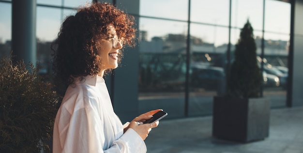 Zijaanzichtfoto van een ondernemer met krullend haar die lacht en een bril draagt tijdens het chatten op de telefoon buiten in de buurt van een modern kantoor
