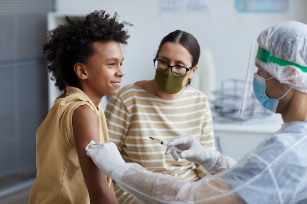 Zijaanzichtportret van een Afro-Amerikaanse tienerjongen die vrolijk glimlacht tijdens covid-vaccinatie in de kliniek, kopieer ruimte