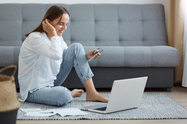 Zijaanzichtportret van een glimlachende mooie vrouw met een wit overhemd en een spijkerbroek die op de vloer zit en naar het display kijkt met een positieve gezichtsuitdrukking die haar haar aanraakt en online betaalt