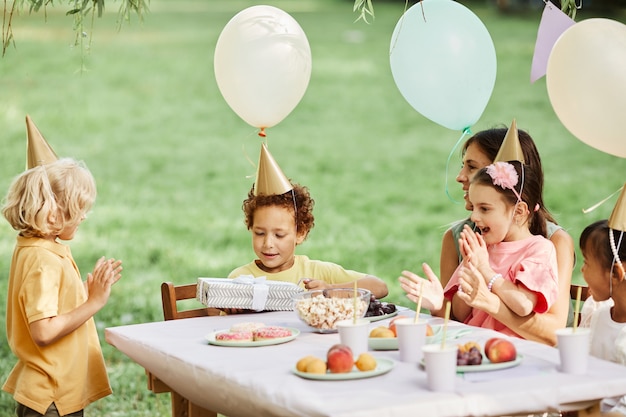 Zijaanzichtportret van een groep kinderen die in de zomer van een verjaardagsfeestje in de buitenlucht genieten en cadeaus geven