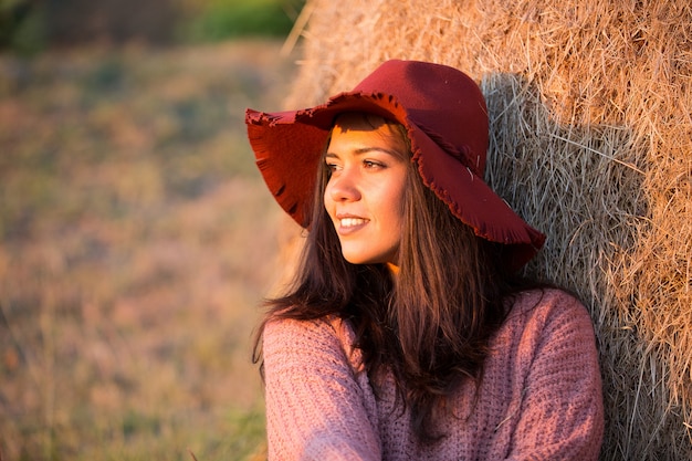 Zijaanzichtportret van mooie jonge vrouw met modieuze hoed bij zonsonderganglicht met wijngaard en strobaal achter haar.