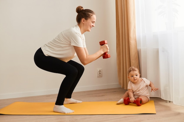 Zijaanzichtportret van slanke atletische vrouw die wit t-shirt draagt dat zich op yogamat bevindt die met dumbbells in handen hurkt en benenuiteinden en armen traint terwijl ze voor de baby zorgt