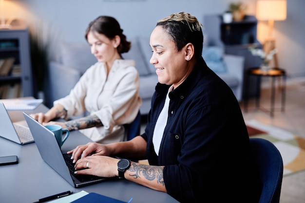 Zijaanzichtportret van twee moderne vrouwen die computers op de thuiswerkplek samen gebruiken en glimlachen