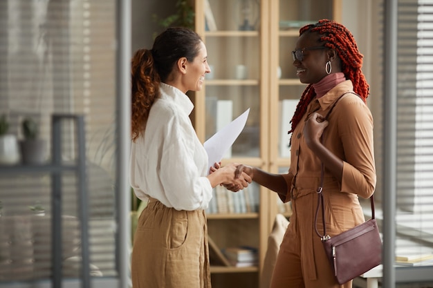 Zijaanzichtportret van twee succesvolle jonge vrouwen die handen schudden en vrolijk glimlachen terwijl status in modern bureaubinnenland, exemplaarruimte