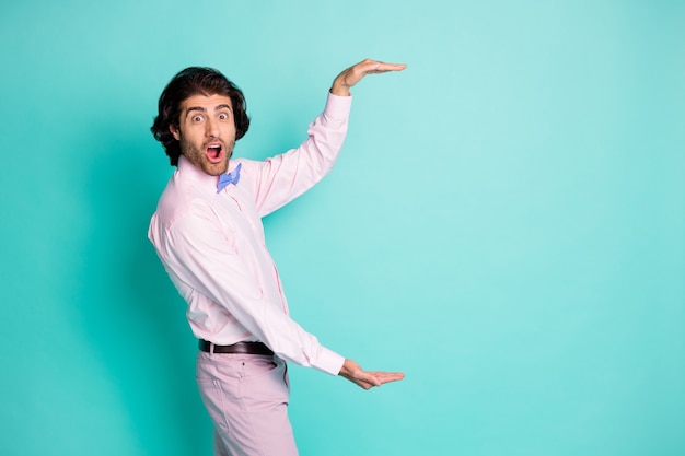 Zijfoto van een vrolijke bruine man met golvend haar die een roze outfit draagt, de lege ruimte meet met twee handen geïsoleerde groenblauwe kleurachtergrond