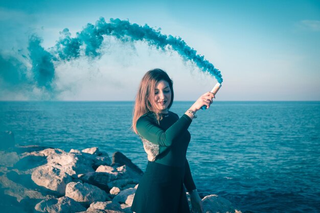 Foto zijkant van een jonge vrouw die aan zee staat met een noodflare tegen een heldere lucht