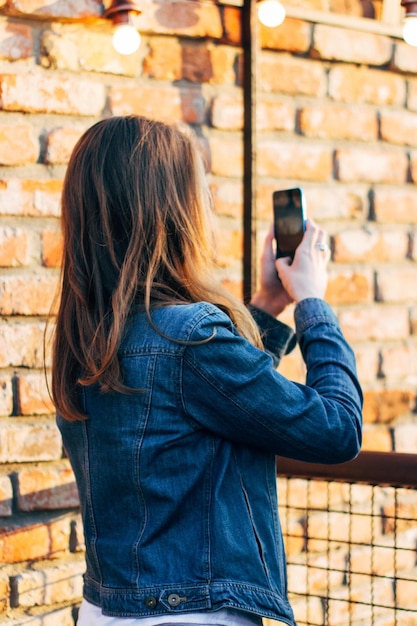 Zijkant van een jonge vrouw die met een mobiele telefoon fotografeert terwijl ze bij een bakstenen muur staat