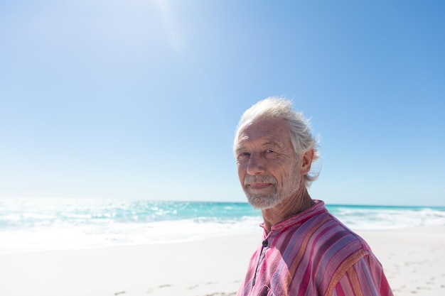 Zijkantbeeld van een oudere blanke man die op het strand staat met blauwe lucht en zee op de achtergrond en naar de camera kijkt