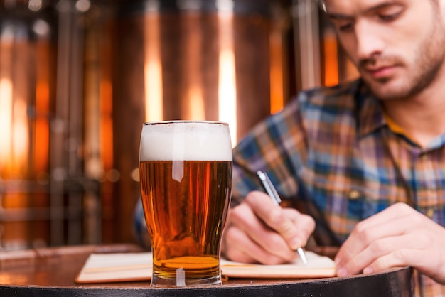 Zijn bierhandel plannen. Zelfverzekerde jonge man in casual shirt die iets in zijn notitieblok schrijft terwijl hij leunt op het houten vat met metalen containers op de achtergrond