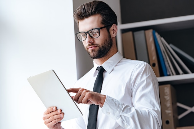 Zijn mail checken. Peinzende jonge knappe zakenman met een bril die zijn touchpad gebruikt terwijl hij op kantoor staat