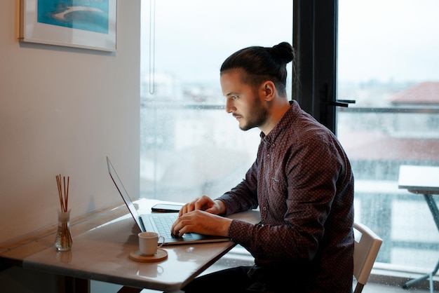 Zijportret van een jonge hipster die thuis aan een laptop werkt op de achtergrond van panoramische ramen