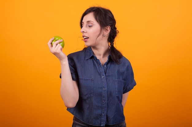 Zijportret van een vrolijke jonge vrouw die een groene appel vasthoudt en bekijkt in de studio op een gele achtergrond. Vrouw die een gezonde levensstijl bevordert. Appels eten voor huidverzorging.