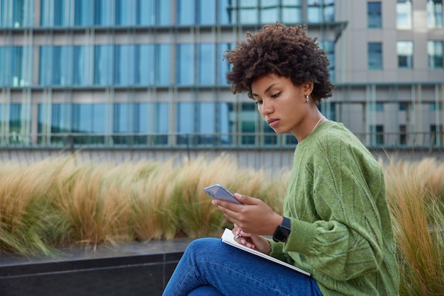 Zijwaarts schot van ernstige vrouw met krullend haar controleert schema online surft informatie op internet via mobiele telefoon schrijft notities in dagboek draagt groene trui en spijkerbroek student kijkt webinar