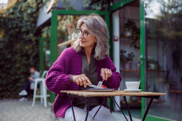 Zilverharige volwassen vrouw in warm gebreid jasje geniet van een heerlijk dessert op het terras van een café