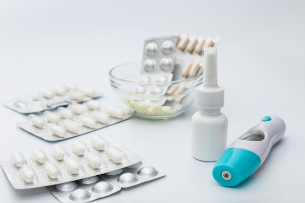 Zilverkleurige blisters met witte capsules en tabletten, een flesje neusspray en een elektronische thermometer