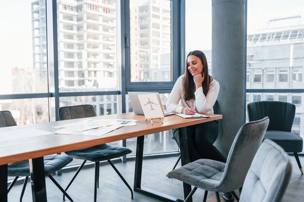 Zit bij de tafel Jonge vrouw in witte formele kleding is binnenshuis in het moderne kantoor