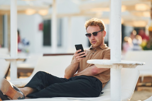 Zit en gebruikt telefoon Jonge man is buiten op zonnige dag Concept van vakantie