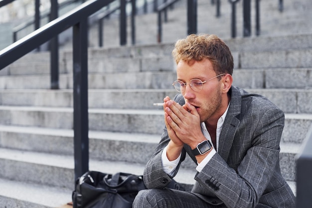 Zit en rookt sigaret Jonge succesvolle zakenman in grijze formele kleding is buiten in de stad