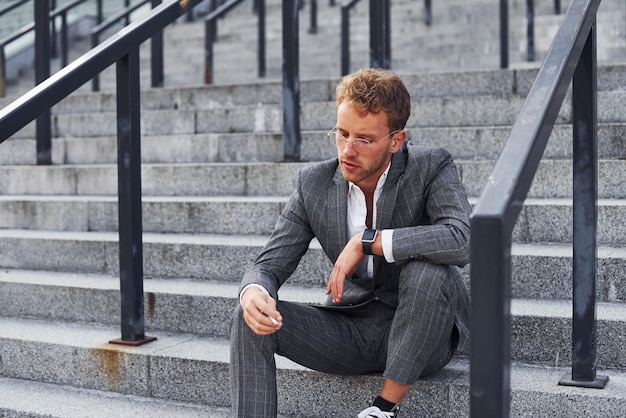 Zit en rookt sigaret Jonge succesvolle zakenman in grijze formele kleding is buiten in de stad