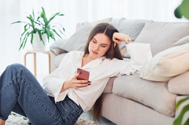 Zit in de buurt van de bank met telefoon in handen Jonge vrouw in wit overhemd en spijkerbroek is thuis