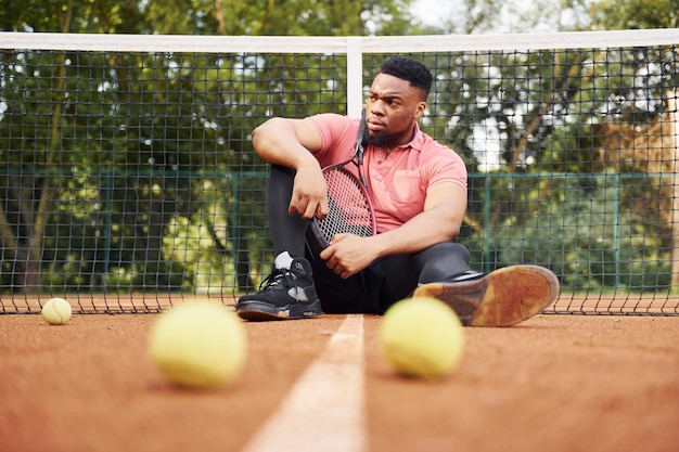 Zit in de buurt van het net en neemt een pauze Afro-Amerikaanse man in roze shirt zit met tennisracket buiten op het veld