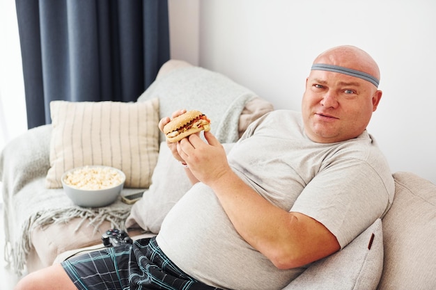 Zit op de bank met popcorn en hamburger Grappige dikke man in vrijetijdskleding is binnenshuis thuis