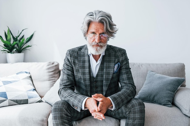 Zit op een comfortabele bank in formele kleding Senior stijlvolle moderne man met grijs haar en baard binnenshuis