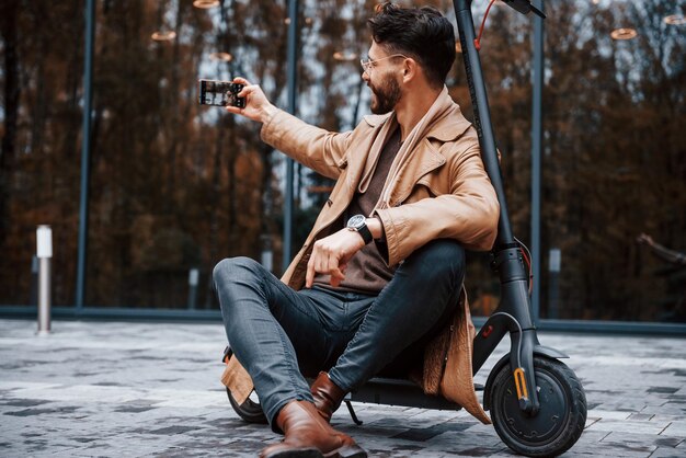 Zit op elektrische scooter Jong mannelijk model in modieuze kleding is overdag buiten in de stad