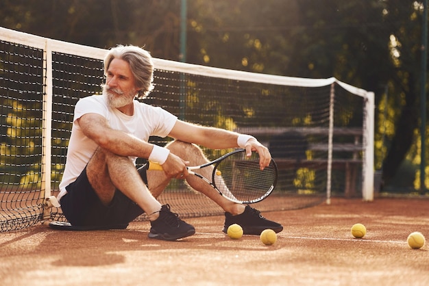 Zitten en een pauze nemen Senior stijlvolle man in wit overhemd en zwarte sportieve korte broek op tennisbaan